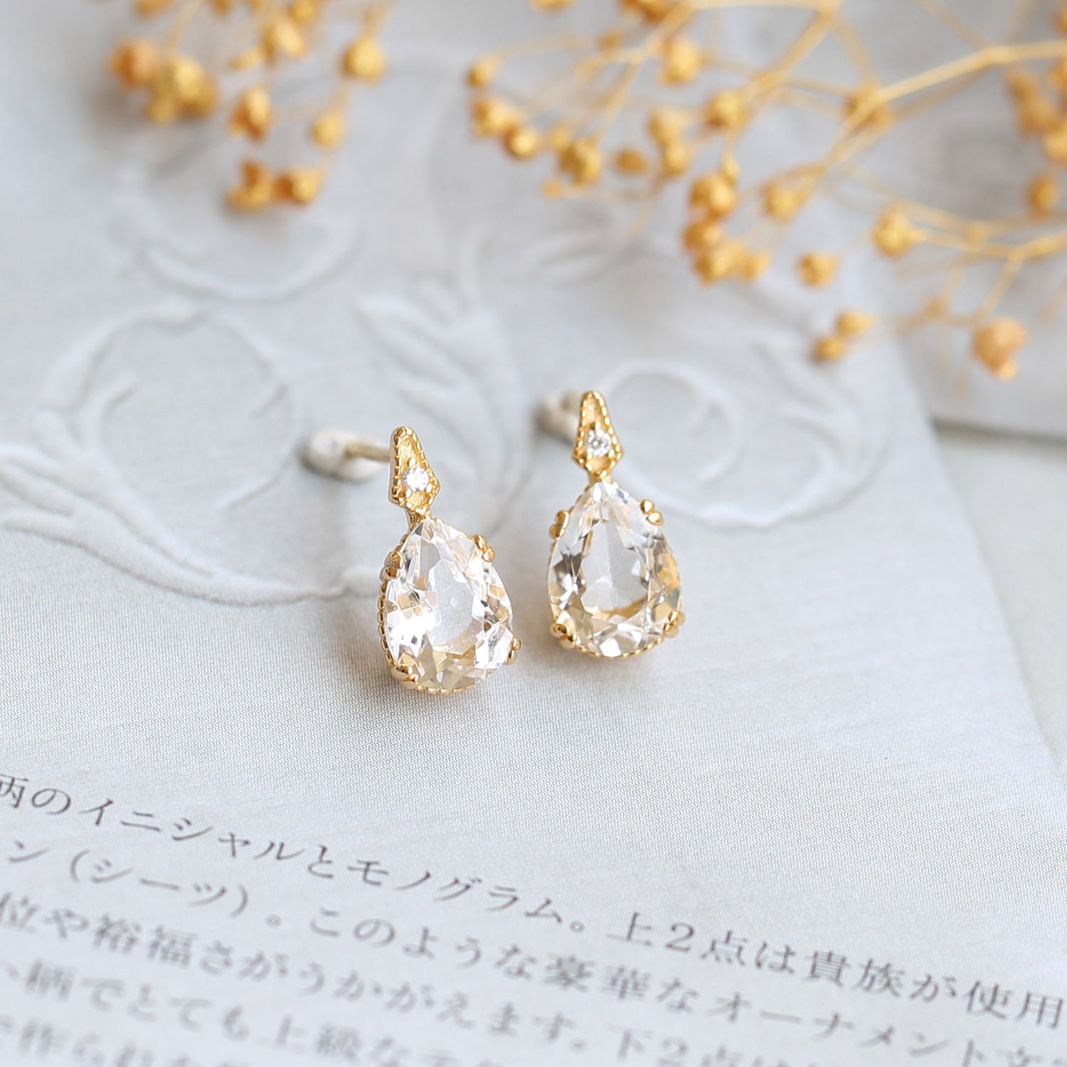 Gold teardrop earrings on a textured surface with dried flowers in the background from the Gembee Jewellery.