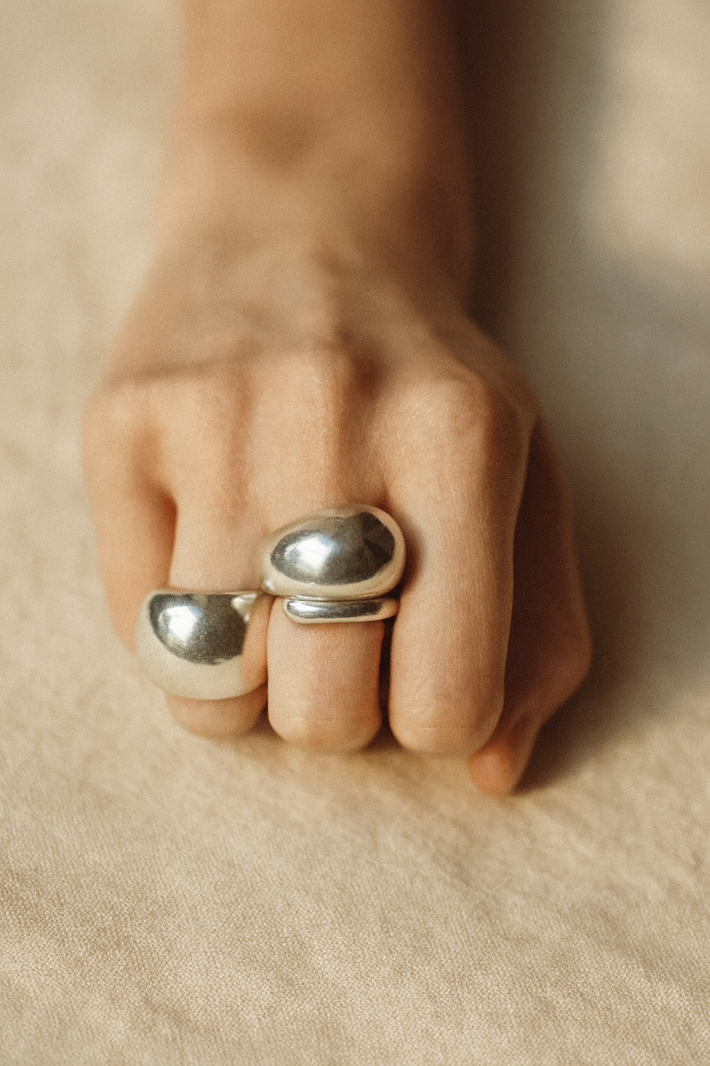 A close-up of a hand wearing two bold, polished silver rings on a neutral fabric background, softly lit to highlight the smooth texture and shine of the jewellery.