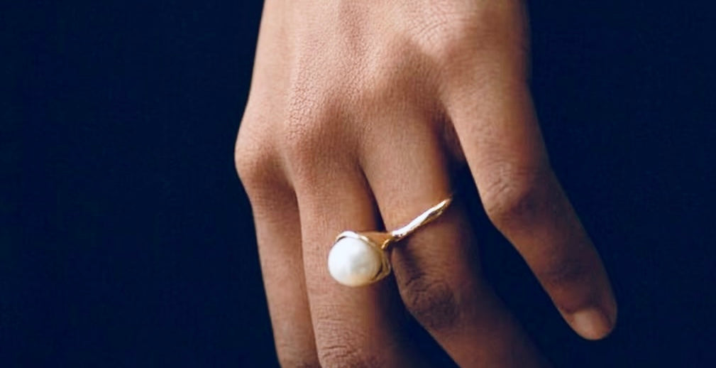 “Close-up of a hand wearing a gold pearl ring with an organic band design against a dark background.