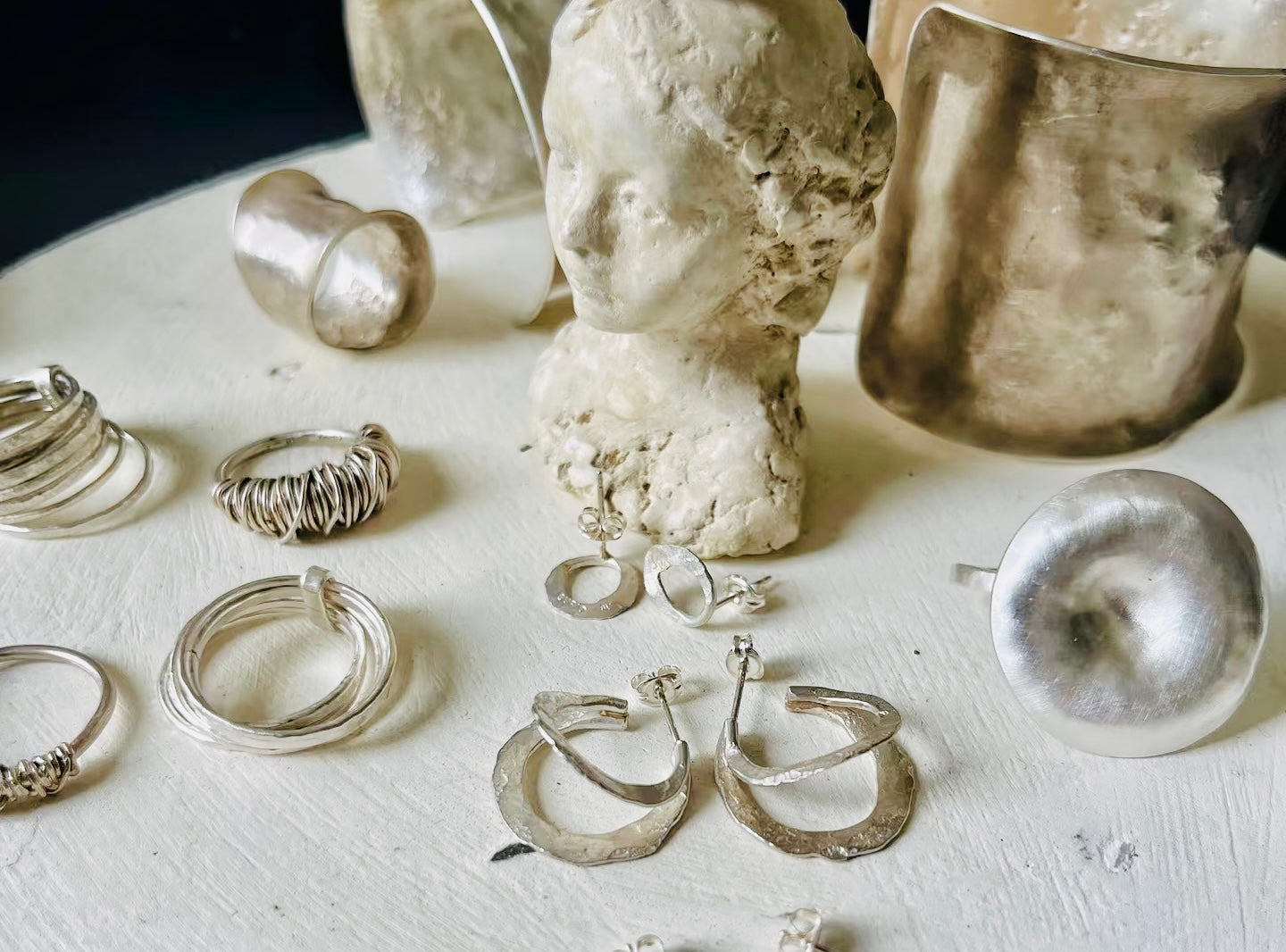 A collection of sterling silver jewellery including textured rings and hoop earrings displayed beside artistic plaster sculptures on a light surface