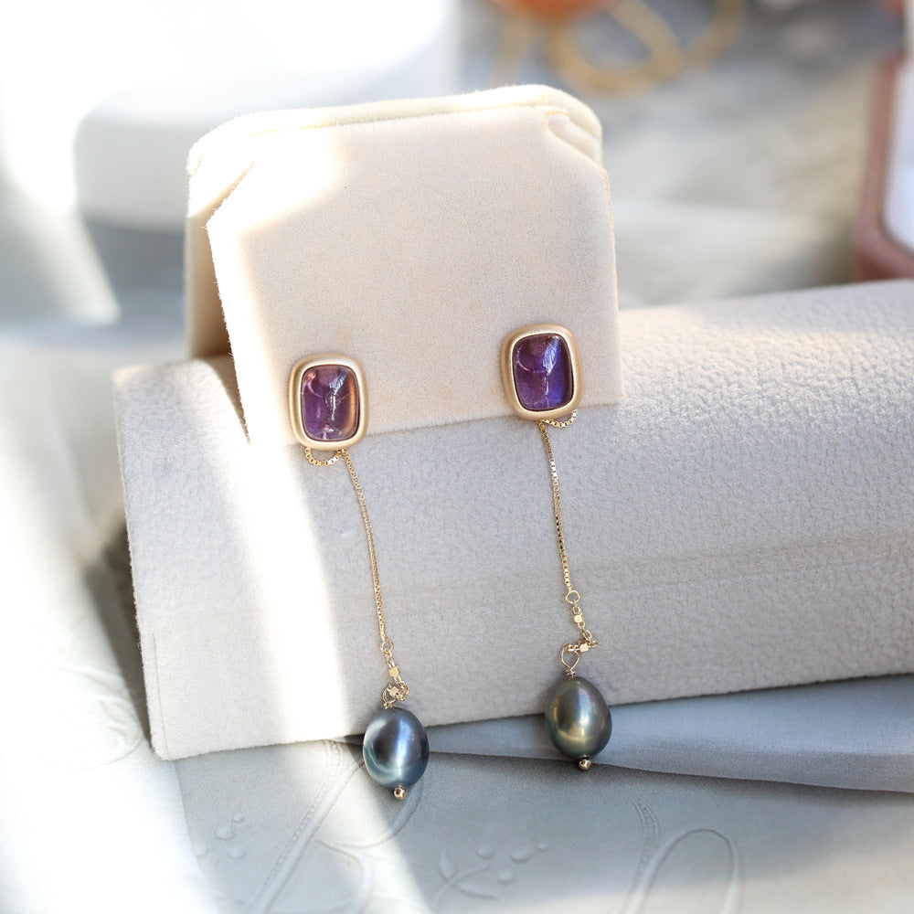 Gold earrings with purple gemstones and black pearls on a white cushion